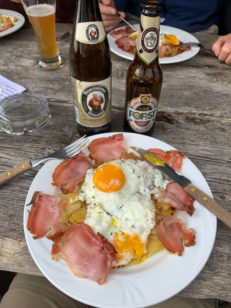 A wooden table with a plate of South Tyrolean food: crispy roasted potatoes topped with two sunny-side-up eggs and slices of cooked ham, served with a Franziskaner Weissbier and a Forst beer, and accompanied by cutlery