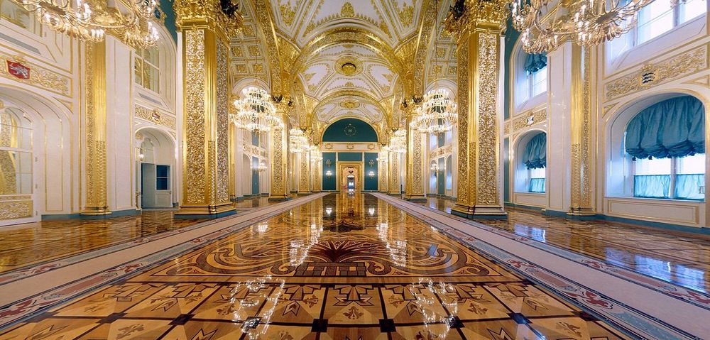 Ornate, grand interior view of Andreevsky Hall in the Grand Kremlin Palace, Moscow. The hall features high vaulted ceilings richly decorated with gold leaf and intricate patterns, massive gilded columns, and sparkling crystal chandeliers. The polished parquet floor reflects the golden light, with elaborate inlaid designs and a central motif. Large arched windows with teal drapery line the walls, and the far end of the hall displays a golden throne beneath a domed arch, symbolizing the hall’s historic role as the imperial throne room dedicated to the Order of St. Andrew the First-Called