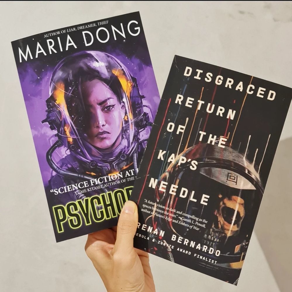 Hand holding two books against a brushed concrete wall: "Disgraced Return of the Kap's Needle" by Renan Bernardo, and "Psychopomp" by Maria Dong.