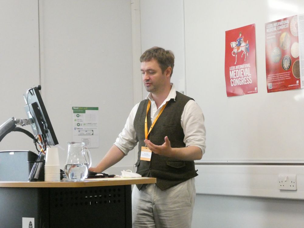 A man with mousy hair, a green-brown waistcoat, and a white shirt looks down at a desk gesturing with his left hand while speaking. A red poster for the International Medieval Congress is behind him on the right.