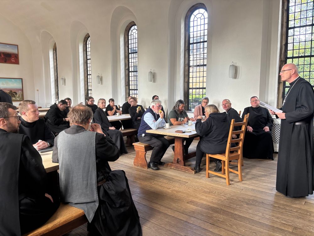 The Principal, a tall man in a cassock, addresses a group of people.