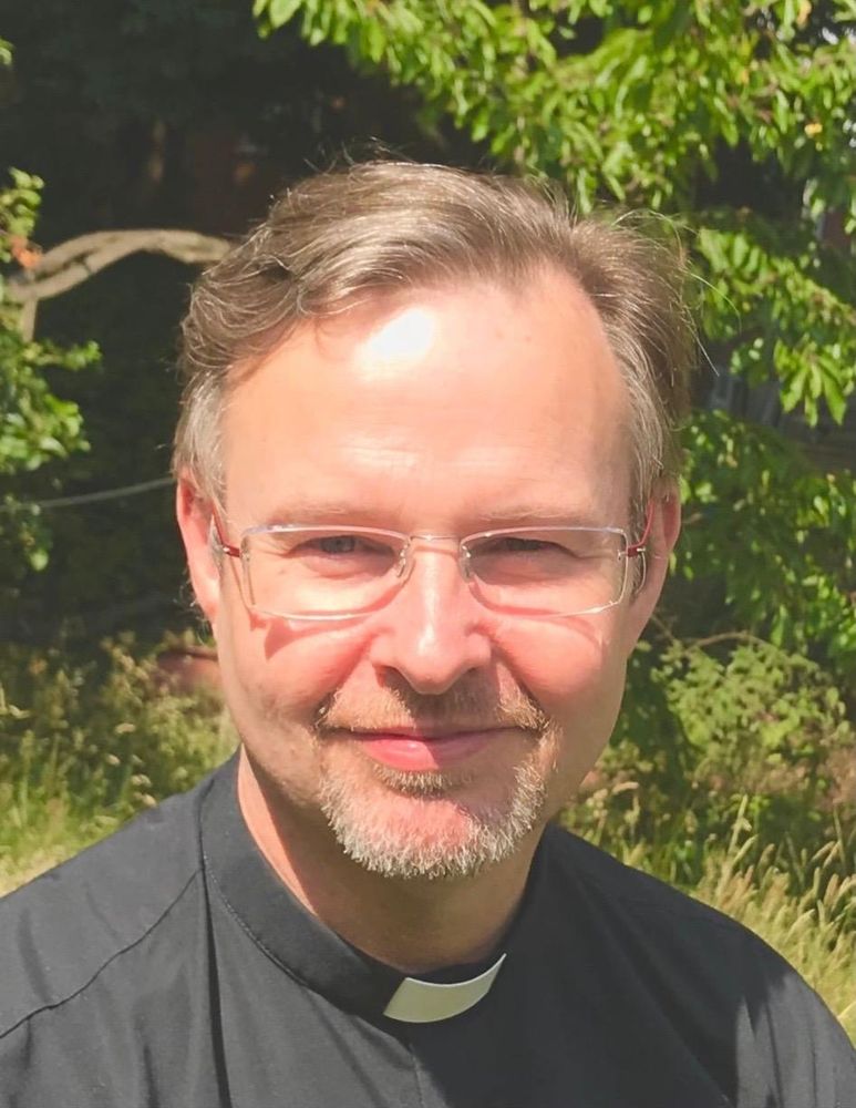A fair haired man with a greying beard in a clerical shirt smiles at the camera.