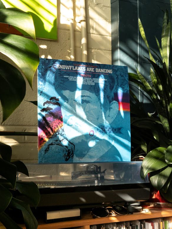 The record sleeve for the album "Snowflakes Are Dancing" by Tomita resting on top of a record player that is spinning that record. It is surrounded by various houseplants and bathed in sunlight.