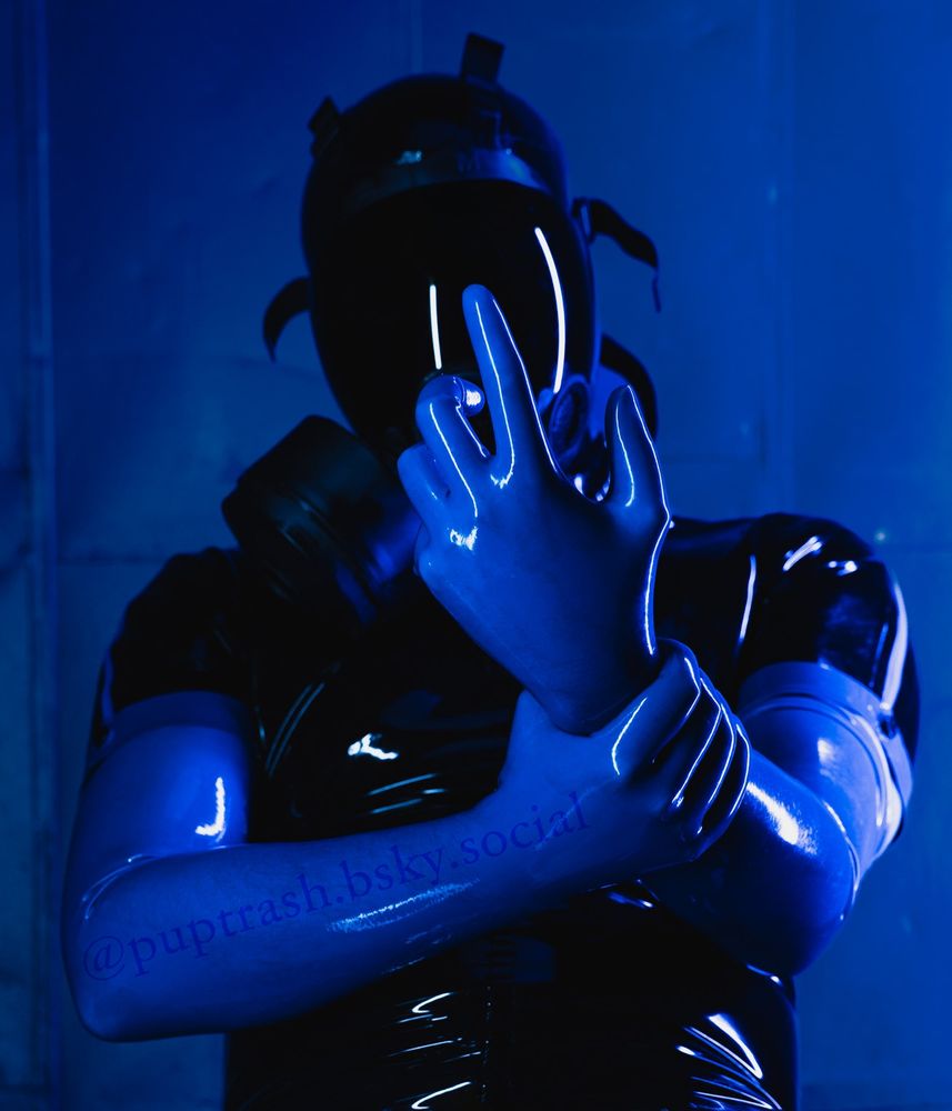 A man in a black latex short sleeve catsuit with blue accents on the sleeves, long blue latex gloves, a black latex hood, and a tinted black MSA gas mask sits in front of a blue metal wall. He holds his left hand out in front of his face and grasps his wrist with his right hand, smoothing out the latex.