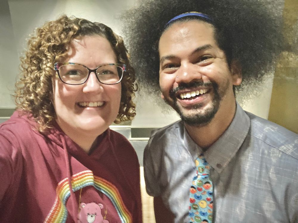 A woman with curly hair wearing a maroon hoodie with a rainbow care bear on it stands happily next to PBS Kids library ambassador and superb human being Mychal Threets.