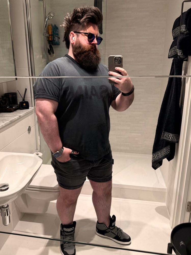 A person with a large beard and styled hair is posing for a mirror selfie in a bathroom. They are wearing sunglasses, a dark gray T-shirt, and denim shorts, paired with high-top sneakers. Various bathroom items and towels are visible in the background
