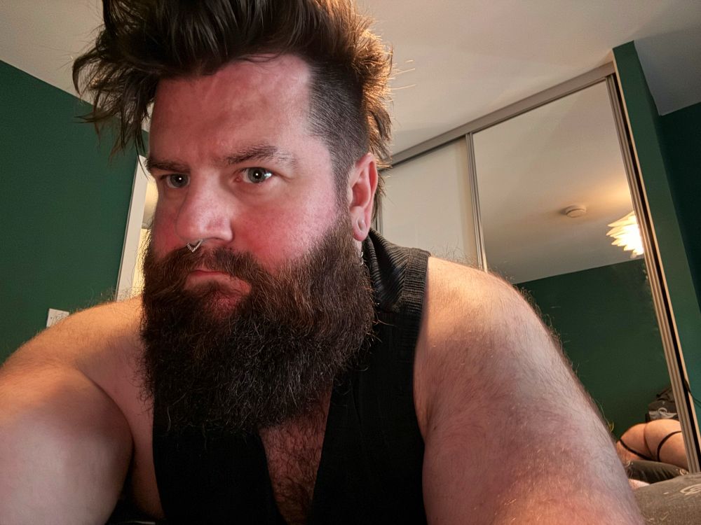 A person with a thick beard and distinct hairstyle is depicted, wearing a black tank top. The background features green walls and a reflection in a mirror. Part of a bed can be seen in the background.