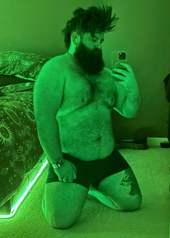 A person with a beard and spiked hair kneeling in a dimly lit room with green lighting. They are shirtless, wearing black shorts, and holding a phone to take a selfie. A bed with patterned bedding and gym equipment is visible in the background 