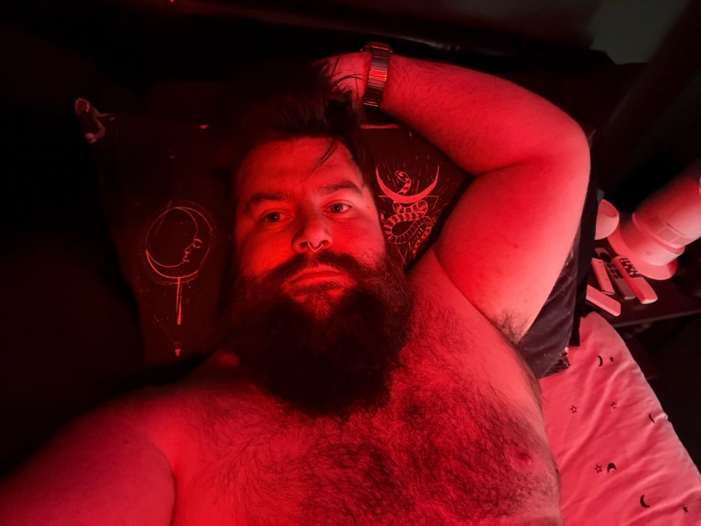 A person with a beard is lying on their back in bed, illuminated by red light. They have their arm resting above their head and are wearing no shirt. A bedside table with various remotes is partially visible.
