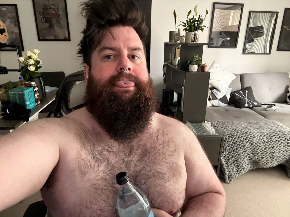 A person with a large beard sits shirtless, sticking out their tongue and holding a water bottle. In the background, there is a monochromatic living room with a couch, decorative pillows, and a table with flowers and various items.