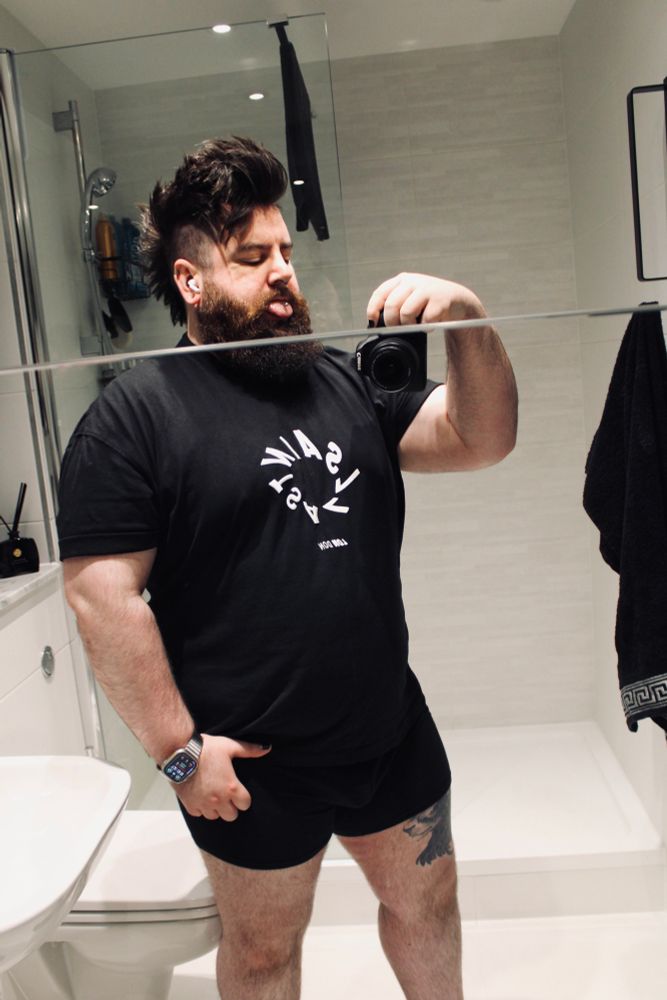 A person with a mohawk hairstyle and a beard is taking a selfie in a bathroom. They are wearing a black t-shirt and black shorts, sticking out their tongue. The bathroom features light colored decor, including a shower and sink.