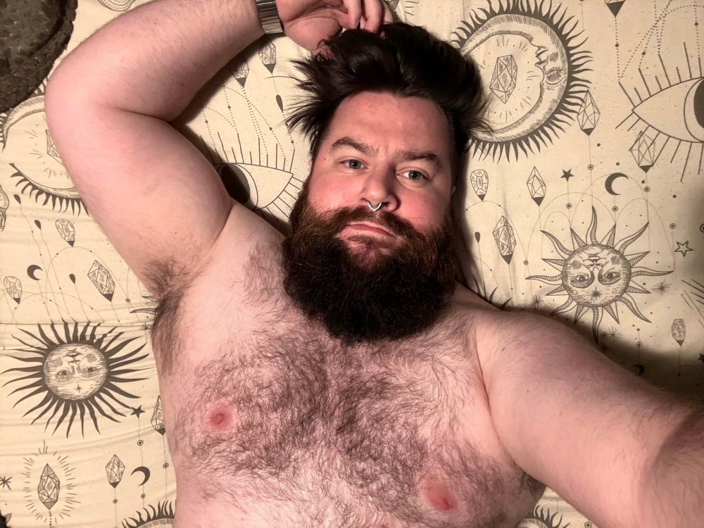 A person with a beard and styled hair is lying on a patterned surface adorned with sun and moon designs. They are shirtless and have a septum piercing. The setting appears casual and relaxed.