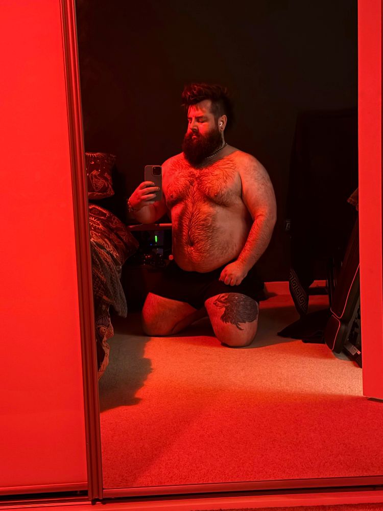A person with a beard is kneeling in front of a mirror, wearing black shorts. The room is dimly lit with red lighting. They are taking a selfie with a smartphone, and there is a tattoo visible on one thigh and a bed with geometric bedding can be seen to one side. 