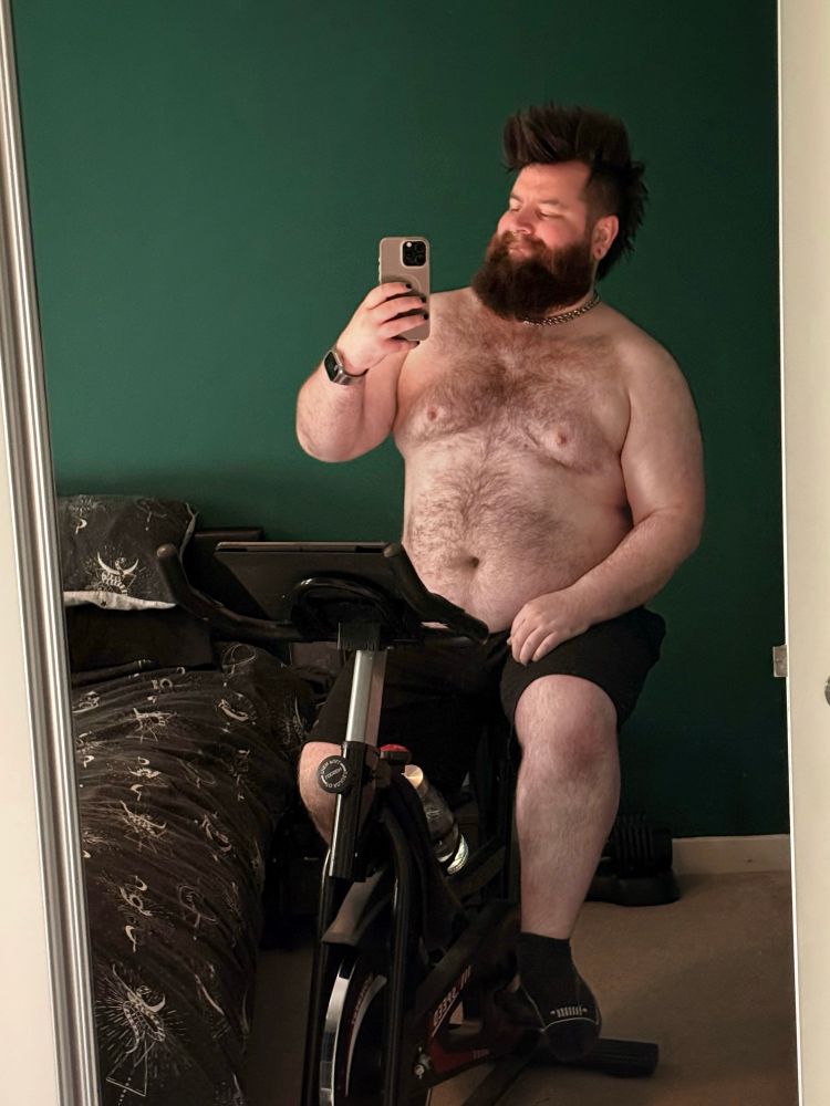 A person with a beard and short hair is sitting on an exercise bike, taking a selfie in a mirror. They are shirtless and wearing shorts, with a bed in the background featuring patterned bedding. The wall behind is painted green.