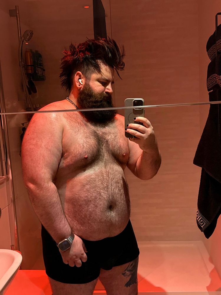 A person with a thick beard and spiky hair taking a selfie in a bathroom. They are shirtless, wearing black shorts, and standing in front of a mirror that reflects a shower and some bathroom items. Soft red lighting is featured. 