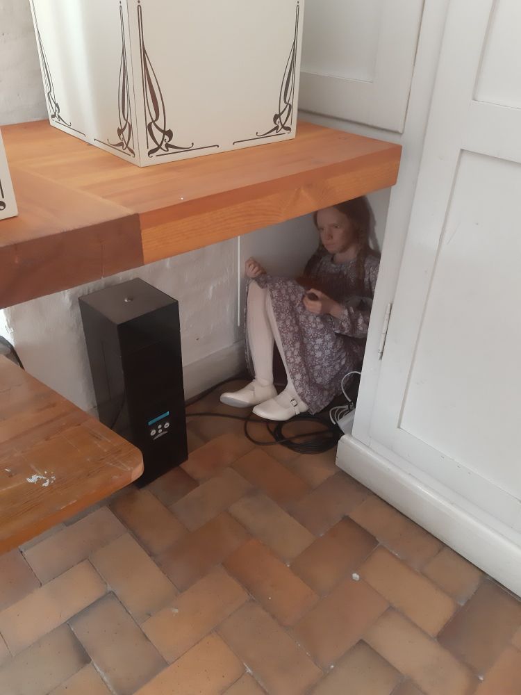 Die lebensechte Puppennachbildung eines Dienstmädchen sitzt zusammengekauert unter einer hölzernen Tischplatte in einer Nische in der Wand. Sie trägt ein einfaches  Kleid mit Blumenmuster und weiße Schuhe. Ihre braunen Haare sind zu zwei Zöpfen geflochten.