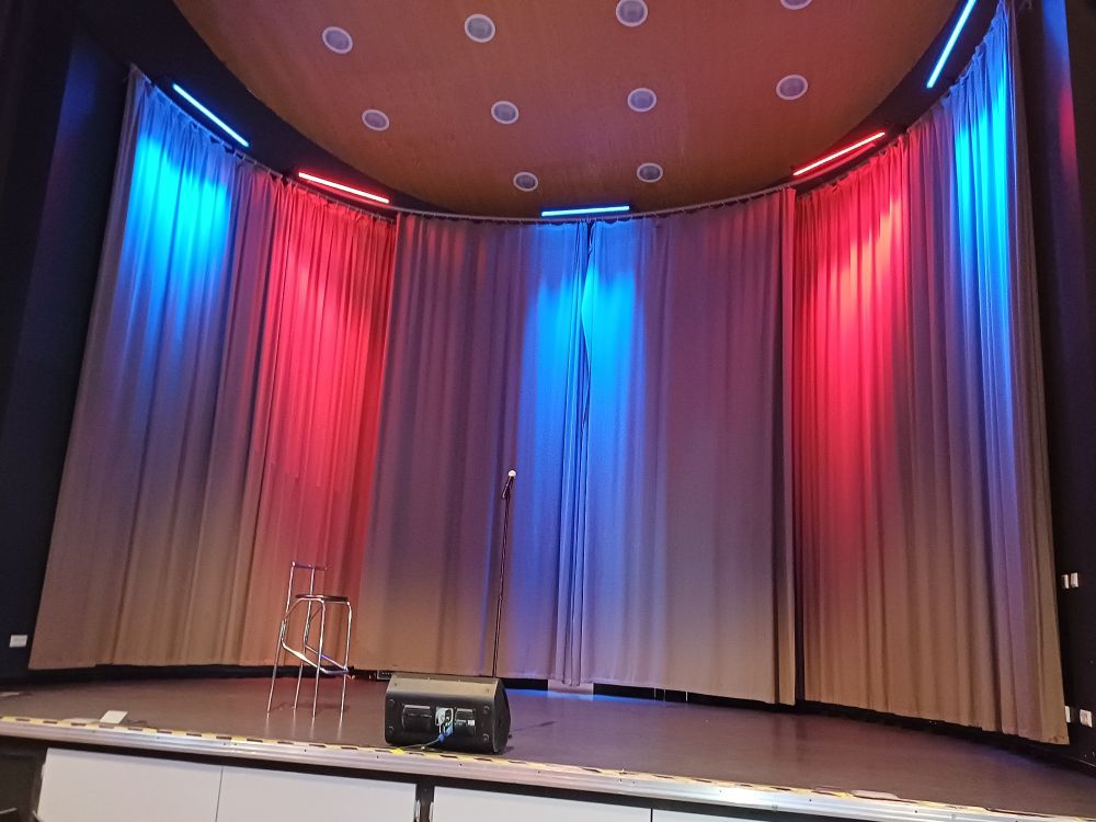 Blick auf eine menschenleere Bühne. Links steht ein Barhocker mit schwarzer Sitzfläche und Metallgestänge.
Der graue Vorhang im Hintergrund wird durch 6 Strahler beleuchtet. Von links nach rechts in blau - rot - blau - rot - blau