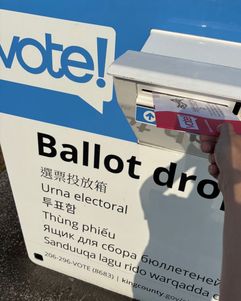 Photo of my hand holding a red-and-white ballot envelope as I slide it into the slot of a ballot drop box that is labeled "vote!" and also says "Ballot drop" in English and several additional languages.