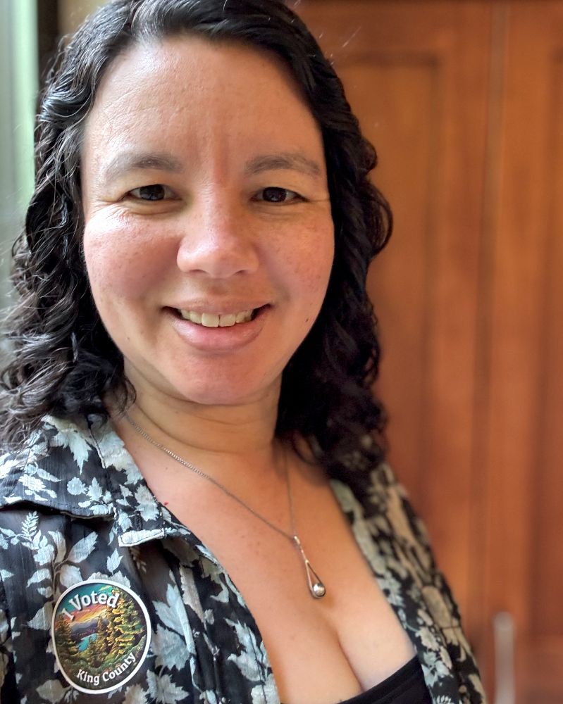 Selfie (me!) a woman with shoulder length curly brown hair, wearing a button-down floral shirt with an "I voted. King County" sticker on it.