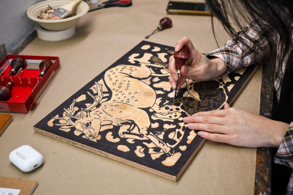 Close-up of Jeni’s hands carving a woodcut. The woodcut imagery is of a Formosan Sika Deer and Clouded Leopard in a cyclical prance / chase, with a branch-like pattern in the background.