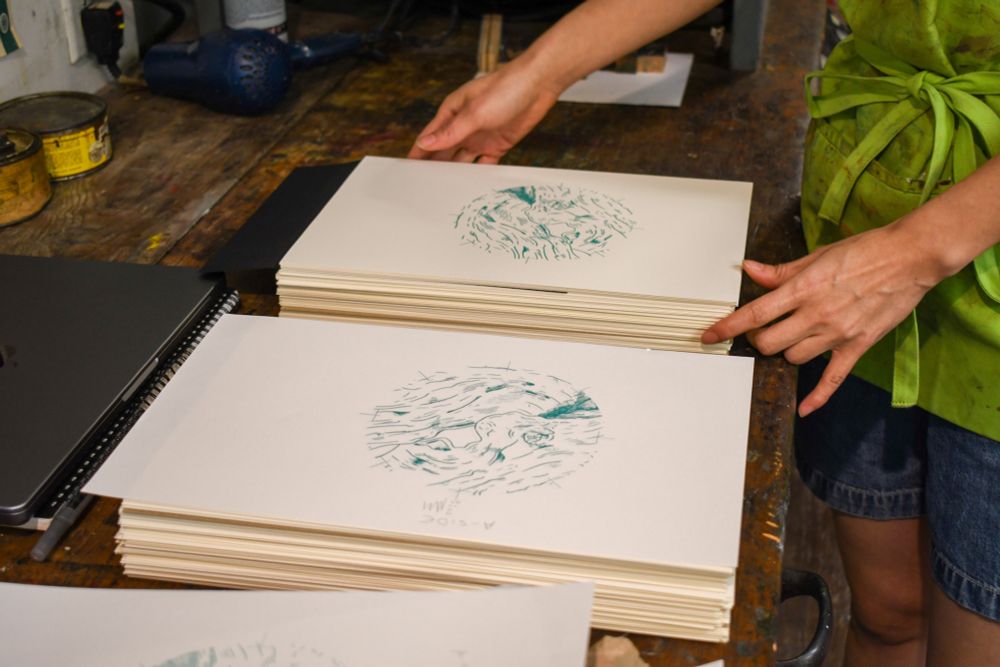 Close-up of Jenie’s hands stacking an edition of prints from the letterpress. The imagery is in teal ink and of a wood grain-like pattern.