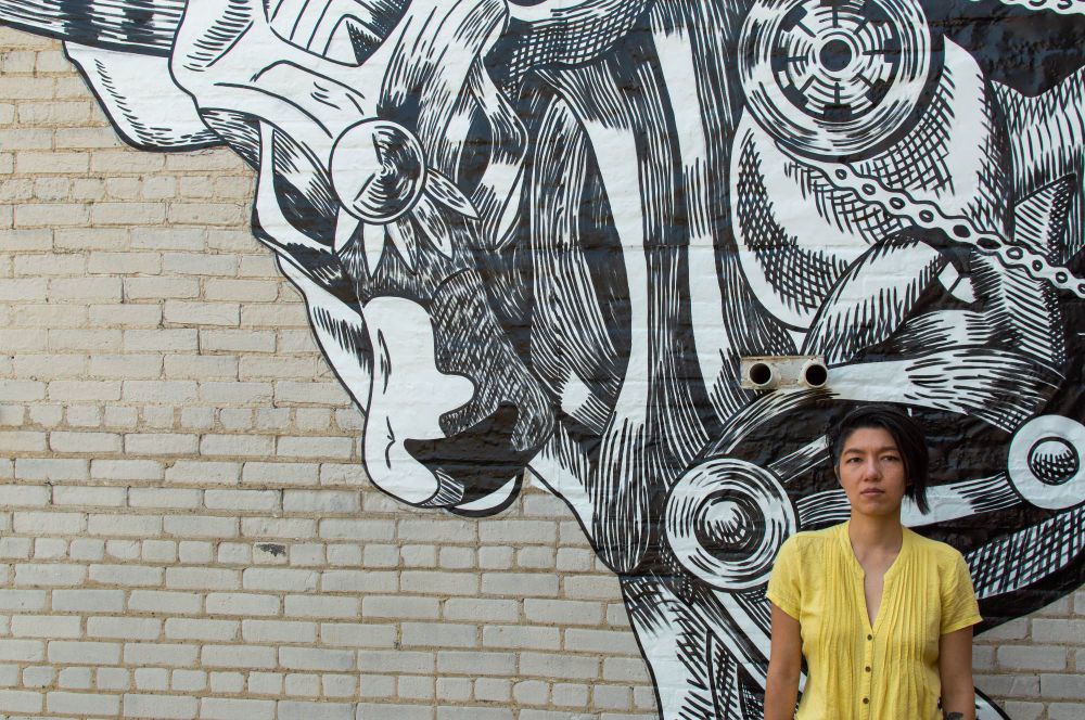 Queer Asian American artist in a yellow shirt in front of a large-scale mural of a steampunk metal ox. The mural is black and white and painted in a cross hatched linework style. This mural is installed in Milwaukee, Wisconsin, and honors the Year of the Metal Ox which brings reliability and resilience. Photo by Adam White Ossers #portfolioday #murals