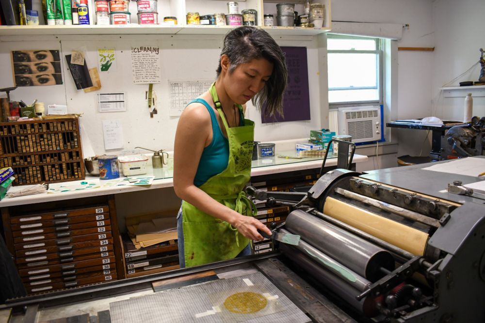 Artist Jenie Gao printing a photopolymer plate on the Vandercook letterpress at Women’s Studio Workshop.

Jenie has short black hair with a side shave and is wearing a teal tank top with a lime green apron.