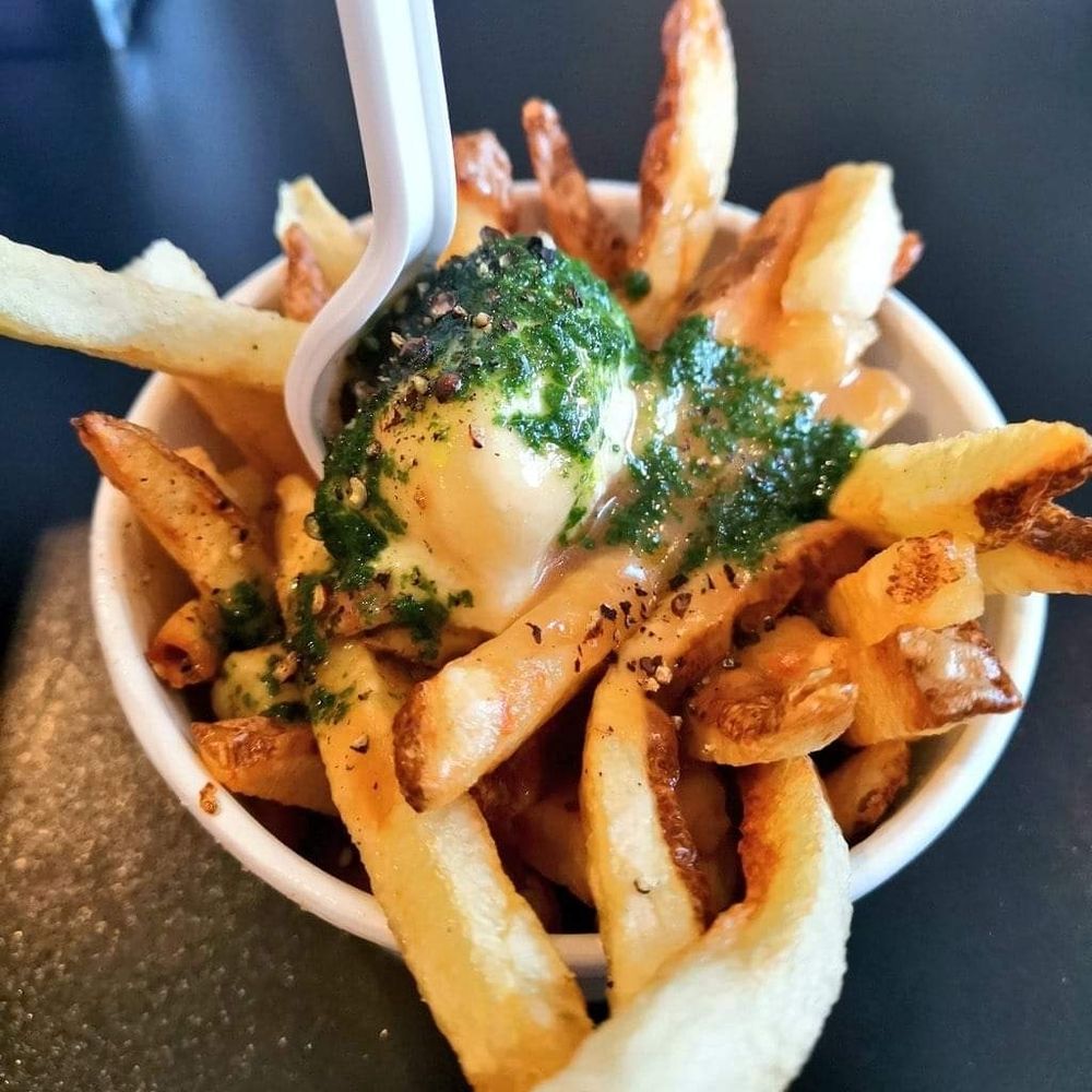 A take out cup filled with hand cut French fries. On top of the fries is a large dollop of goat's cheese drizzled with a fresh basil oil and fresh cracked black pepper. The cheese is starting to melt from the hot gravy that is poured over it. A goat's cheese and basil poutine.