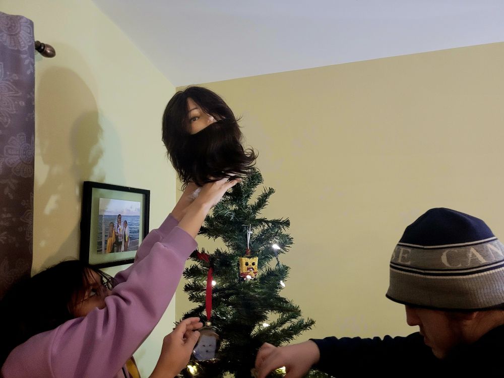 My kids decorating our tree. One is putting a hairstyle modeling head on the top. The head has long hair and a beard.