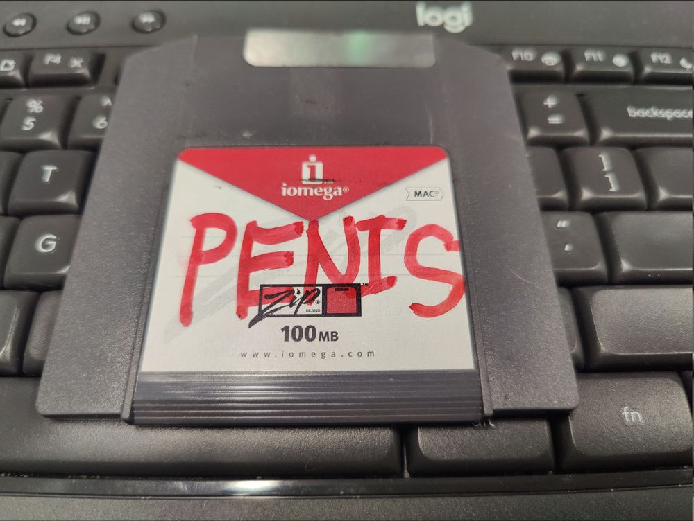 100MB Zip 100 disk, sitting on a logitech wireless keyboard as a photo backdrop with the word "PENIS" written across the label in red permanent marker.