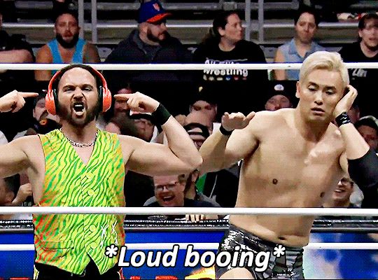 okada on the right miming DJing and scratching record, nick jackson is on the left with large headphones on, pointing at them while mouthing "i can't hear you". the caption on the gif is "Loud booing"