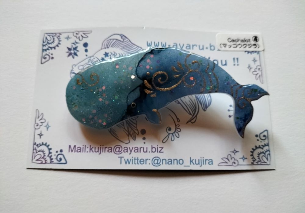 Une broche bleue avec dorures, représentant un cachalot, par @nano_kujira sur Twitter.