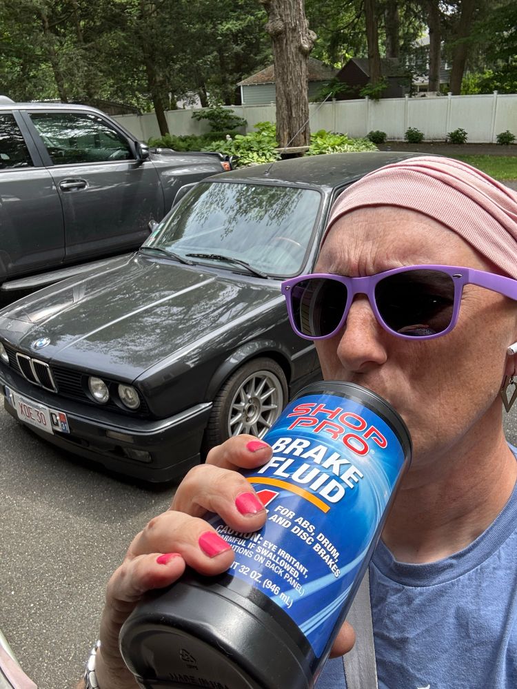 Person in purple glasses standing in front of 1989 BMW E30 touring (wagon). They appear to be drinking from a giant bottle of DOT 4 Brake Fluid. Visible on the label is the warning “Caution: Eye irritant, harmful if swallowed.”