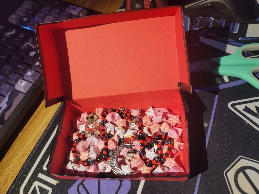 A red and black beaded necklace with a large moon charm in a red and black giftbox. There are also paper stars and a small figure of an alien with a keytar