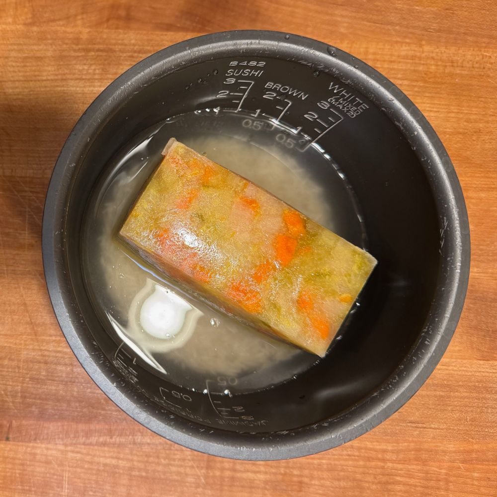 A rice cooker's bowl, containing some rice, water, and (most importantly) a block of frozen mirepoix. 