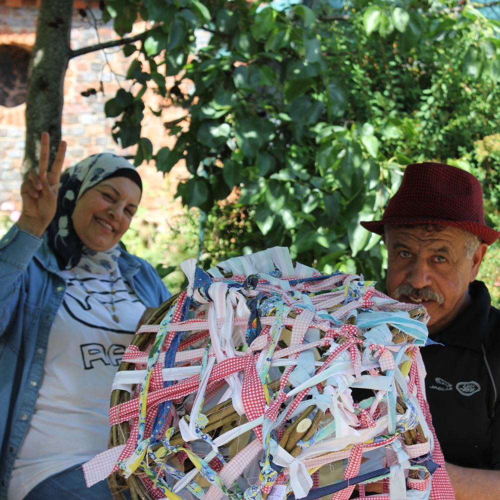 Two people working on a community weaving project. One, a woman in a headscarf, poses for the photo, holding two fingers up in a peace sign