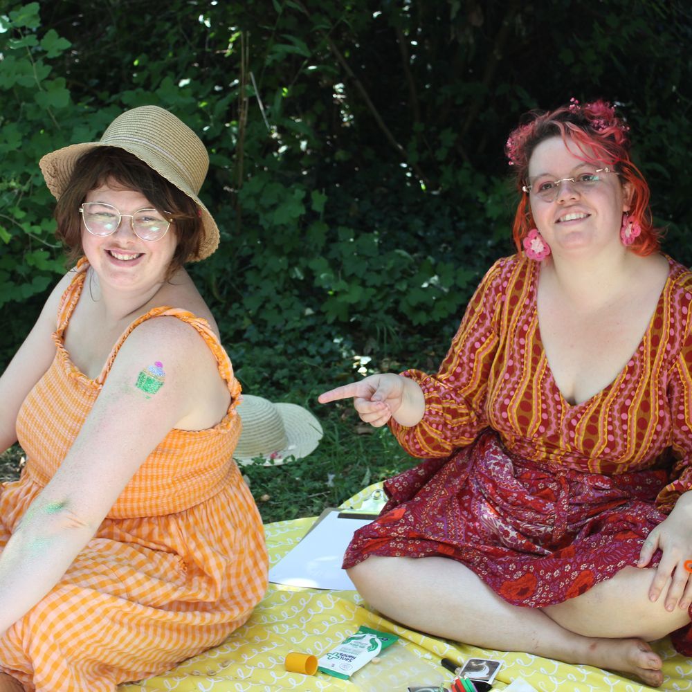 Two people sitting on a picnic blanket in colourful outfits. One points to the other's glitter tattoo