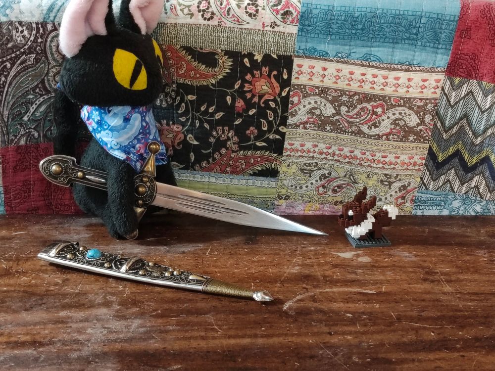Miss L cat plush wearing the "snow foxes" scarf holding an unsheathed, ornate dagger, pointing it at a nanoblock Eevee