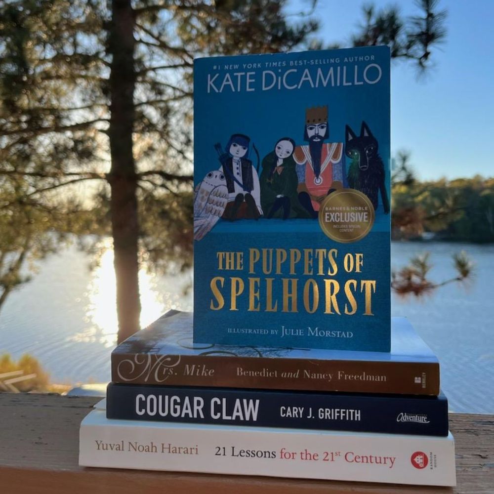 Four books: Kate DiCamillo's "The Puppets of Spelhorst", Benedict and Nancy Freedman's "Mrs. Mike", Cary J. Griffith’s “Cougar Claw”, and Yuval Noah Harari’s "21 Lessons for the 21st Century”. Books are displayed on an outdoor porch railing. They are visible against the backdrop of a shadowy pine tree and the sun setting behind a glistening lake.