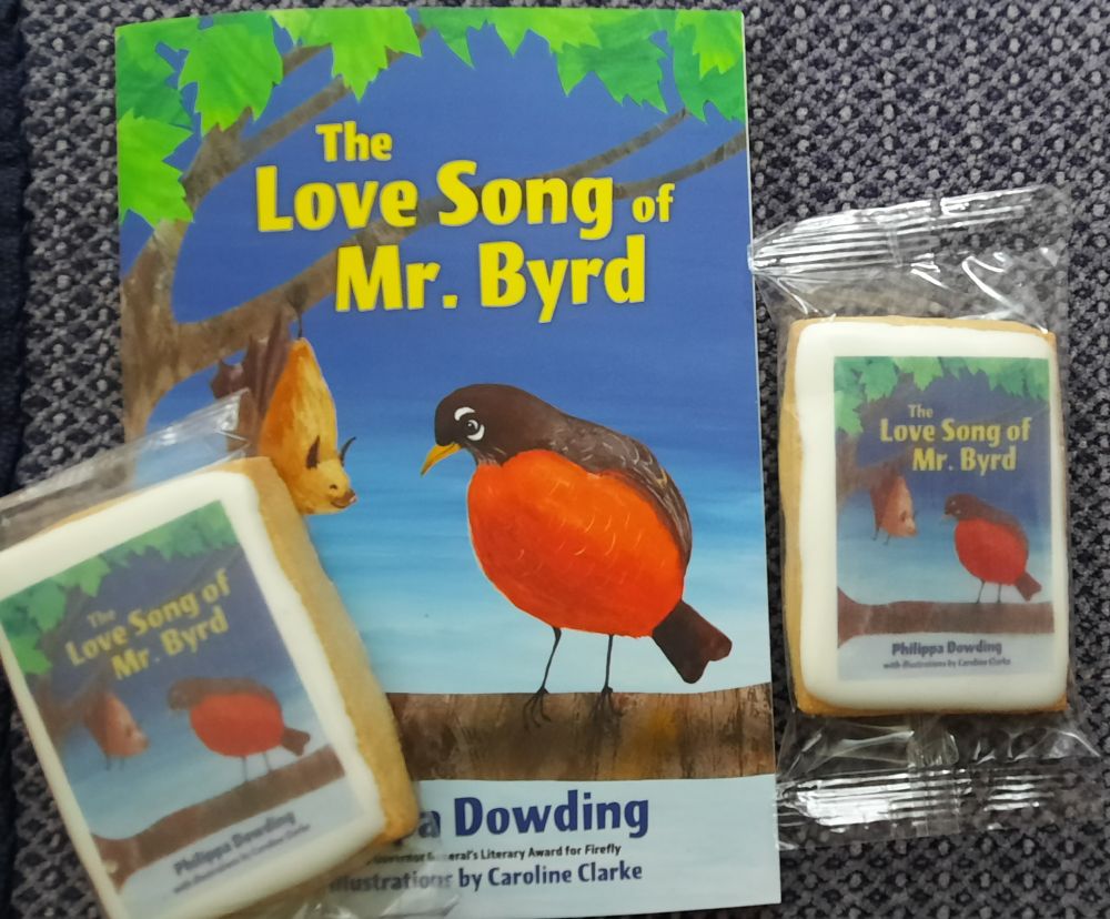 "The Love Song of Mr. Byrd", a new chapter book by Philippa Dowding. The image shows the book cover with bird and bat on a tree limb looking at each other. One shortbread "book cookie" with the cover imprinted on it in icing is placed on either side.