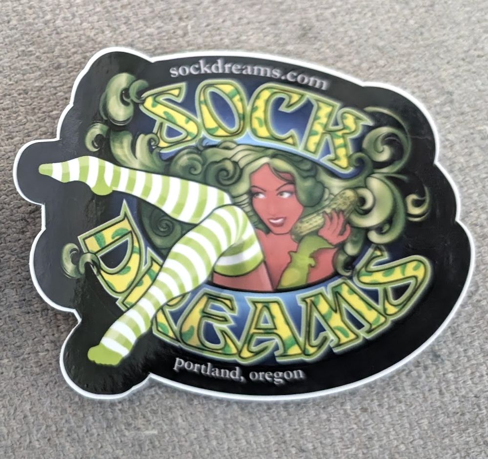 Sock Dreams (Portland, OR) sticker, pickle themed, green and yellow and blue. The babe in their logo has green hair and is holding a very large pickle