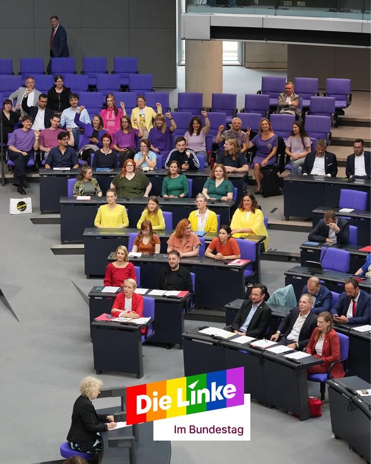 Die Abgeordenten der Fraktion Die Linke sitzen auf ihren Plätzen im Plenum. Sie sind, Reihe für Reihe, bunt gekleidet, sodass sich aus ihren Outfits eine Regenbogenflagge ergibt.