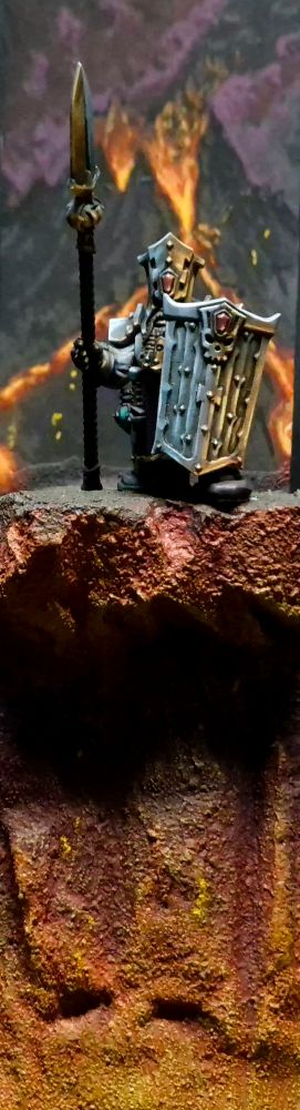 A Chaos Dwarf stands on the edge of a lava lake with a volcano erupting off in the distance.