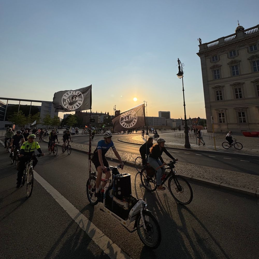Das Bild zeigt eine Gruppe von Fahrradfahrern, die in einer Stadt, möglicherweise Berlin, an einer Demonstration oder Fahrradparade teilnehmen.  Der Sonnenuntergang beleuchtet die Szene.  Die Fahrräder fahren auf einer Straße vor historischen Gebäuden.  Einige Teilnehmer tragen Fahnen mit dem Aufdruck "Respect Cyclists"  und dem Bild einer Evolution des Radfahrens.  Ein Polizeibeamter auf dem Fahrrad ist ebenfalls sichtbar.  Das Bild vermittelt ein Gefühl von Gemeinschaft und Aktivismus im Zusammenhang mit dem Radfahren.
