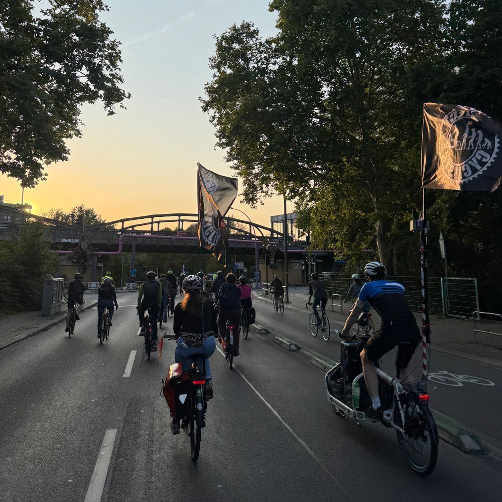 Das Bild zeigt eine Gruppe von Radfahrern, die auf einer Straße fahren. Die Sonne geht unter, und der Himmel ist orange und rosa. Die Radfahrer tragen Fahnen mit einem Logo darauf, das wie ein Radfahrersymbol aussieht.  Im Hintergrund ist eine Brücke zu sehen. Das Gesamtbild wirkt friedlich und vermittelt den Eindruck einer gemeinsamen Radtour oder einer Demonstration.
