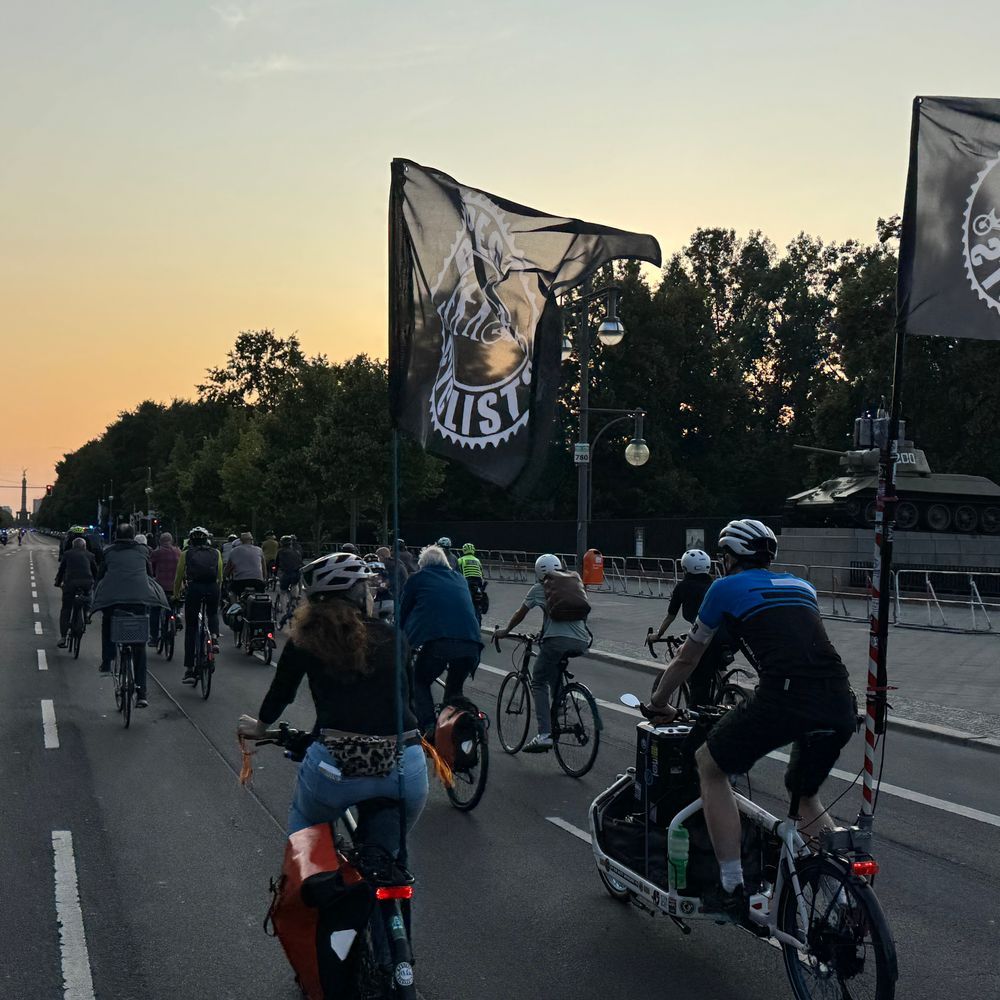 Das Bild zeigt eine Gruppe von Radfahrern, die in einer Reihe auf einer Straße fahren. Im Hintergrund ist ein Panzer zu sehen, und am Himmel ist ein Sonnenuntergang zu beobachten. An der Spitze der Fahrradkolonne wehen zwei Fahnen, eine davon trägt ein Rad-Motiv und die Aufschrift „RESPECT CYCLISTS“. Die Szene scheint eine Fahrraddemonstration oder -kundgebung zu sein, die möglicherweise gegen den Militarismus oder für den Radverkehr gerichtet ist.  Die Stimmung ist eher ruhig und ernst.
