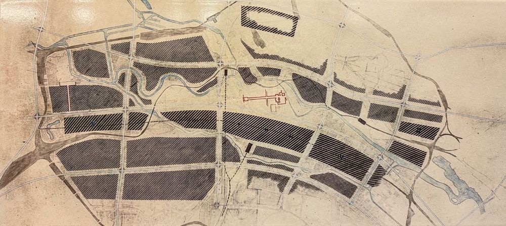 Das Bild zeigt einen Stadtplan oder eine städtebauliche Planungsskizze. Es sind rechteckige, schraffierte Flächen zu sehen, die wahrscheinlich Baublöcke oder größere Areale darstellen. Dazwischen verlaufen Straßen und Flüsse. In der Mitte des Plans ist ein komplexes, rot gezeichnetes Gebäude oder eine Anlage zu erkennen. Der Zeichenstil wirkt handgefertigt und erinnert an Entwürfe aus einer früheren Epoche.