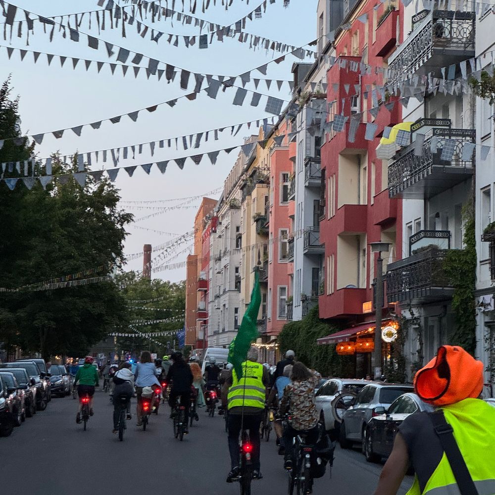 Das Bild zeigt eine Fahrradgruppe, die am Abend eine Straße entlangfährt. Die Straße ist von mehrstöckigen Häusern mit Balkonen gesäumt, die mit Wimpeln geschmückt sind. Ein Fahrradfahrer trägt eine grüne Fahne. Die Stimmung ist ruhig und friedlich.  Im Hintergrund ist ein Schornstein zu erkennen.
