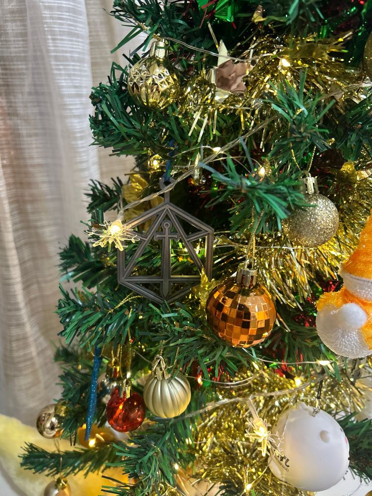 A tight shot of a metal Critical Role logo Christmas tree ornament, next to a glittery white bear Christmas ornament 