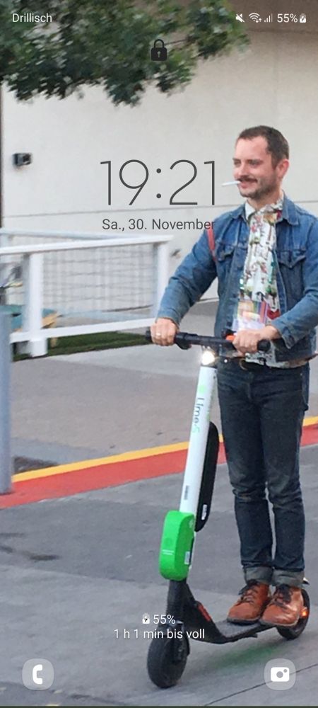 Screenshot von meinem Sperrbildschirm. Hinter den gängigen Icons wie Uhrzeit, Datum und Kamera, ist ein Bild von Elijah Wood auf einem E-Scooter. Er trägt braune Lederschuhe, eine dunkle Jeans, ein buntes Hemd und eine Jeansjacke. Er hat eine Zigarette im Mund und sieht extrem zufrieden aus mit sich und der Welt.